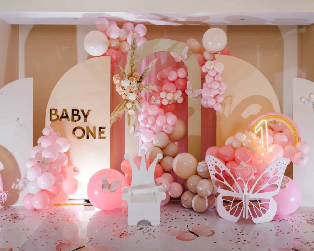 Arch decorated pink balloons, rainbow, text baby one, flowers, paper decor butterfly, and wooden white chair. Birthday party for 1 year old girl on a background photo wall. Children's photo zone.